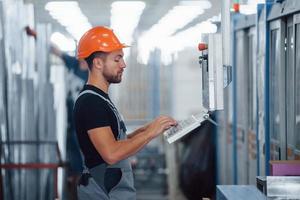 Using machine to program operations. Industrial worker indoors in factory. Young technician with orange hard hat photo
