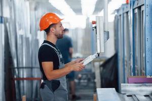 Using machine to program operations. Industrial worker indoors in factory. Young technician with orange hard hat photo