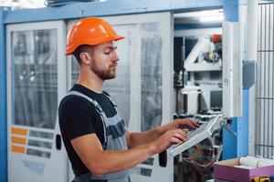 Using machine to program operations. Industrial worker indoors in factory. Young technician with orange hard hat photo
