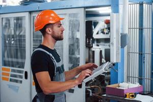 Using machine to program operations. Industrial worker indoors in factory. Young technician with orange hard hat photo