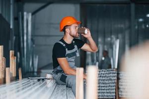 With cup of drink. Lunch break. Industrial worker indoors in factory. Young technician with orange hard hat photo