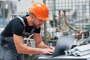 portátil moderno. trabajador industrial en el interior de la fábrica. joven técnico con casco naranja foto