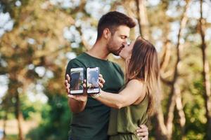 Holding two smartphones with photos of them. Beautiful young couple have a good time in the forest at daytime