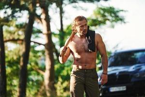 Holds wooden axe. Handsome shirtless man with muscular body type is in the forest at daytime photo