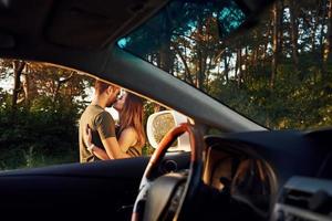 View from the car's interior. Steering wheel, side mirror. Beautiful young couple have a good time in the forest at daytime photo