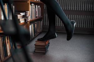 Standing on books and trying to reach top of shelves. Female student is in library. Conception of education photo