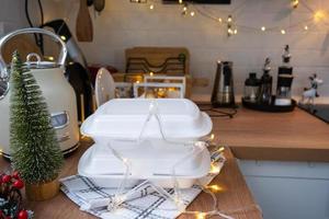 Food delivery service containers on table white scandi festive kitchen in christmas decor. Eve New year, saving time, too lazy to cook, hot order, disposable plastic box in fairy light. mock up photo