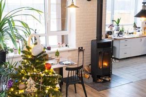 Festive interior of house is decorated for Christmas and New Year in loft style with black stove, fireplace, Christmas tree. Warm studio room with white kitchen, burning wood, cozy and heating of home photo