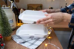 Food delivery service containers on table white scandi festive kitchen in christmas decor. Eve New year, saving time, too lazy to cook, hot order, disposable plastic box in fairy light. mock up photo