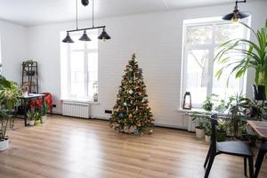 Christmas tree in the white interior of a house with large windows. Glowing fairy lights garlands interior decoration of the studio room. Potted plants in the home photo