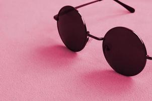 Stylish black sunglasses with round glasses lies on a blanket made of soft and fluffy fleece fabric. Image toned in Viva Magenta, color of the 2023 year photo