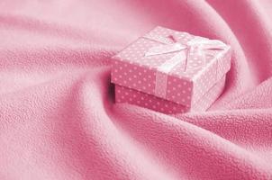 A small gift box with a small bow lies on a blanket of fleece fabric with a lot of relief folds. Packing for a gift to your lovely girlfriend Image toned in Viva Magenta, color of the 2023 year photo