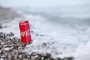 antalya, turquía - 18 de mayo de 2022 la lata roja original de coca cola se encuentra sobre pequeñas piedras redondas cerca de la orilla del mar. coca-cola en playa turca foto