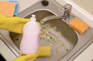 el limpiador muestra una botella de detergente líquido en el fregadero de la cocina sucia con partículas de comida antes de la limpieza