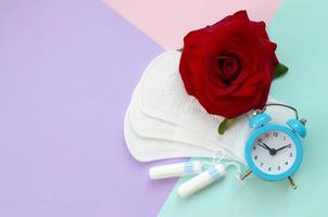 Menstrual pads and tampons with blue alarm clock and red rose flower on multicolored background photo