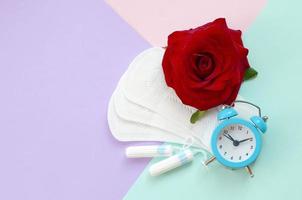 Menstrual pads and tampons with blue alarm clock and red rose flower on multicolored background photo