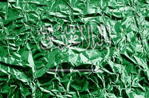 Saudi Arabia flag depicted in paint colors on shiny crumpled aluminium foil closeup. Textured banner on rough background photo