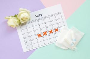 Flat lay composition with white roses and menstrual tampons and pad packs on menstruation period calendar and blue pink and lilac pastel background photo