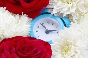 Little cute blue alarm clock surrounded by red roses and chrysanthemum heads photo