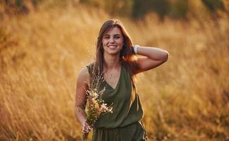 una chica hermosa camina por el campo con hierba alta y recogiendo flores. increíble luz del sol foto