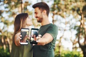 sosteniendo dos teléfonos inteligentes con fotos de ellos. hermosa pareja joven diviértete en el bosque durante el día