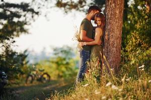 Leaning on the tree. Beautiful young couple have a good time in the forest at daytime photo