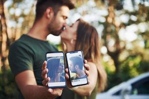 Holding two smartphones with photos of them. Beautiful young couple have a good time in the forest at daytime