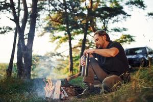 Making drink by using thermos. Man in black shirt near the campfire in the forest at his weekend time photo