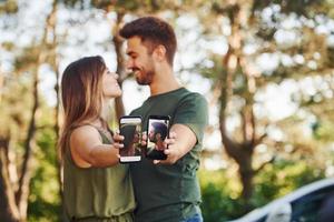 Holding two smartphones with photos of them. Beautiful young couple have a good time in the forest at daytime