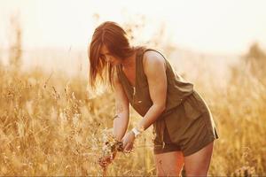 Beautful girl walks through the field with high grass and collecting flowers. Amazing sunlight photo