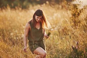 Beautful girl walks through the field with high grass and collecting flowers. Amazing sunlight photo