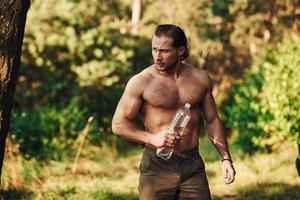 With bottle of water. Handsome shirtless man with muscular body type is in the forest at daytime photo