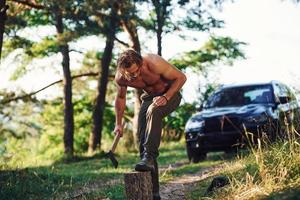 leñador con un hacha cortando madera. un hombre guapo sin camisa con un tipo de cuerpo musculoso está en el bosque durante el día foto