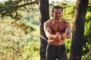 Doing workout. Handsome shirtless man with muscular body type is in the forest at daytime photo