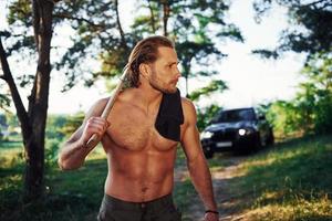 Close up portrait of woodsman with axe in hand. Handsome shirtless man with muscular body type is in the forest at daytime photo