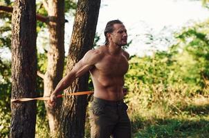 Doing workout. Handsome shirtless man with muscular body type is in the forest at daytime photo