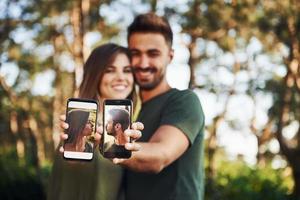 sosteniendo dos teléfonos inteligentes con fotos de ellos. hermosa pareja joven diviértete en el bosque durante el día