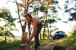 leñador con un hacha cortando madera. un hombre guapo sin camisa con un tipo de cuerpo musculoso está en el bosque durante el día foto