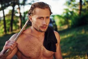 Close up portrait of woodsman with axe in hand. Handsome shirtless man with muscular body type is in the forest at daytime photo