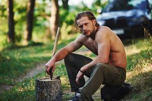 Holds wooden axe. Handsome shirtless man with muscular body type is in the forest at daytime photo