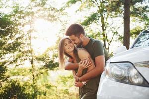 Front part of white car. Beautiful young couple have a good time in the forest at daytime photo