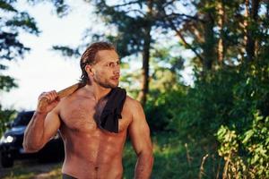 Close up portrait of woodsman with axe in hand. Handsome shirtless man with muscular body type is in the forest at daytime photo