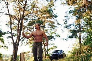 Woodsman with an axe cutting wood. Handsome shirtless man with muscular body type is in the forest at daytime photo