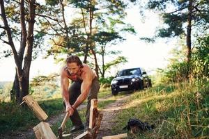 Woodsman with an axe cutting wood. Handsome shirtless man with muscular body type is in the forest at daytime photo