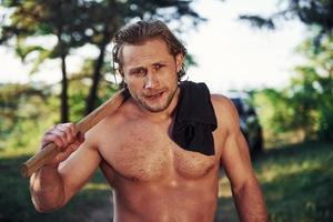 Close up portrait of woodsman with axe in hand. Handsome shirtless man with muscular body type is in the forest at daytime photo