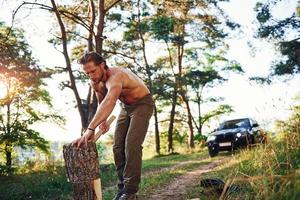 Woodsman with an axe cutting wood. Handsome shirtless man with muscular body type is in the forest at daytime photo