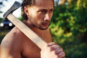 Confident look. Close up portrait of woodsman with axe in hand. Handsome shirtless man with muscular body type is in the forest at daytime photo