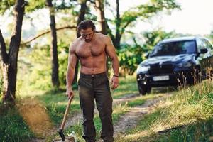 Holds wooden axe. Handsome shirtless man with muscular body type is in the forest at daytime photo