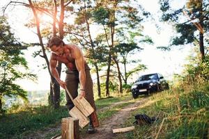 Woodsman with an axe cutting wood. Handsome shirtless man with muscular body type is in the forest at daytime photo