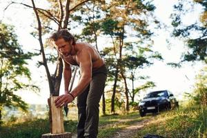 leñador con un hacha cortando madera. un hombre guapo sin camisa con un tipo de cuerpo musculoso está en el bosque durante el día foto